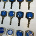 May include: Close-up view of sixteen vintage keys with blue circular inserts. Each insert displays a unique number, such as 202, 206, 272, 302, 1007, 1110, 1520, 1210, 2154, 2166, 2172, and 2617. The keys are made of brass or a similar metal, showing signs of age and wear. The blue inserts are slightly recessed into the key's body. The text 'THIS SIDE UP' is visible on some keys. These are likely hotel room keys or similar vintage numbered keys.