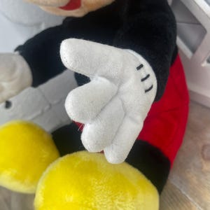 May include: A plush Mickey Mouse toy with black fur, red shorts, and white gloves. The toy is sitting with its hands in front of it.