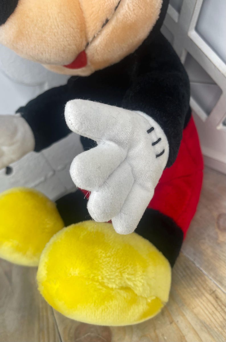 May include: A plush Mickey Mouse toy with black fur, red shorts, and white gloves. The toy is sitting with its hands in front of it.