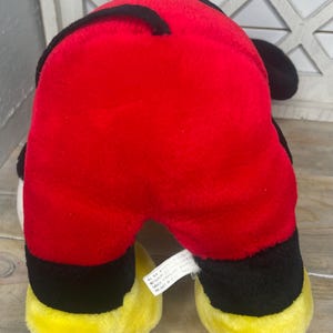 May include: Red, black and yellow plush toy of a cartoon dog. The toy has a tag on the bottom that reads "All New Material. Made in China. Reg. No. PA-11543 (CN) CA24145-17000. Product of China."