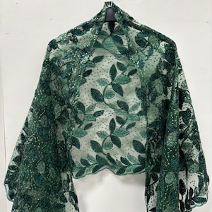 Puede incluir: Un chal transparente de color verde esmeralda con un delicado estampado floral de hojas. El chal está hecho de una tela ligera, con un borde festoneado. El diseño presenta un patrón repetido de hojas y pequeños puntos.