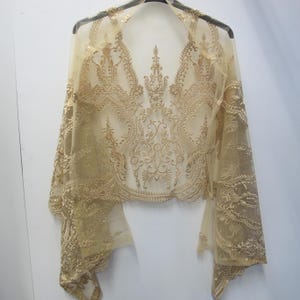 Puede incluir: Un chal de encaje transparente de color dorado con intrincados bordados florales y de volutas. El chal está drapeado sobre una percha, mostrando su delicado diseño fluido. Las mangas son anchas y los bordes están festoneados.