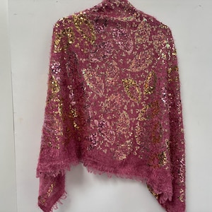 Op de afbeelding: Een roze sjaal met gouden en zilveren pailletten bladpatronen. De sjaal heeft een fuzzy textuur en een gefranjerde rand. Het kledingstuk hangt tegen een witte achtergrond.