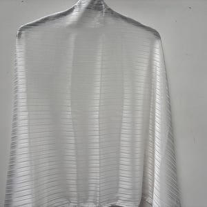 Puede incluir: Una tela blanca texturizada drapeada sobre una percha. La tela tiene un sutil patrón de rayas horizontales. El material parece ligero y fluido, lo que sugiere que podría ser una bufanda o un chal.
