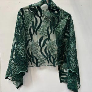 Op de afbeelding: Een donker smaragdgroene bolero gemaakt van doorschijnende mesh-stof met ingewikkelde bloemenborduursels en pailletten. Het kledingstuk heeft lange mouwen en een geschulpte zoom, met een gedetailleerd patroon van bladeren en bloemen.