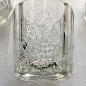 Puede incluir: Un vaso de vidrio transparente con un diseño grabado de uvas y hojas. El vaso tiene una forma cuadrada y un fondo redondeado.