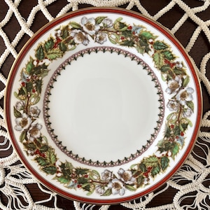 May include: White ceramic plate with a red border and gold trim. The plate features a floral wreath design with white flowers, green leaves, and red berries. The plate is likely for decorative or serving purposes.