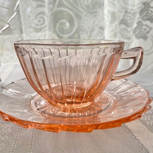 May include: A pink glass teacup and saucer set with a fluted design. The cup has a handle and the saucer has a scalloped edge.