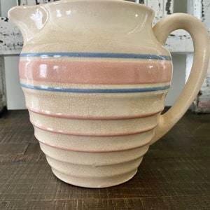 May include: A vintage cream-colored ceramic pitcher with a pink and blue striped design. The pitcher has a textured, ribbed surface and a handle.