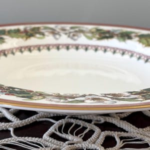 Puede incluir: Un cuenco de cerámica blanca con un borde decorativo. El borde presenta un borde rojo con un ribete dorado y un patrón de hojas verdes y bayas rojas. El cuenco está sobre un tapete de encaje blanco.