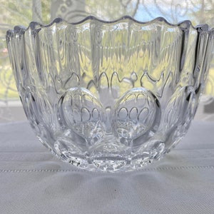 Puede incluir: Un cuenco de vidrio transparente con un borde festoneado y un diseño texturizado de círculos en relieve. El cuenco está vacío y se encuentra sobre un mantel blanco.