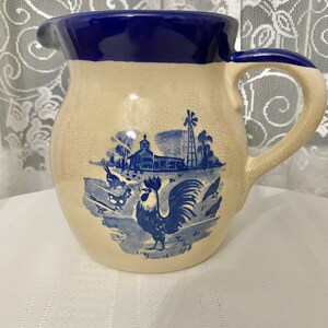 Puede incluir: Una jarra de cerámica azul y blanca con un gallo y una escena de granja. La jarra tiene un borde y un asa azules.
