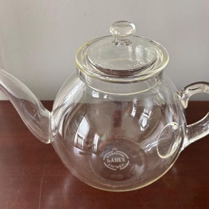 Puede incluir: Una tetera de vidrio transparente con un cuerpo redondeado, un pico curvo y un asa. La tapa también es de vidrio transparente y tiene un pequeño pomo. La tetera tiene una etiqueta en la parte inferior que dice "Pyrex".