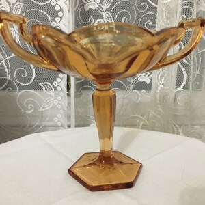 以下が含まれることがあります： 2つのハンドルと六角形のベースを持つ、アンバーガラスのビンテージキャンディーディッシュです。