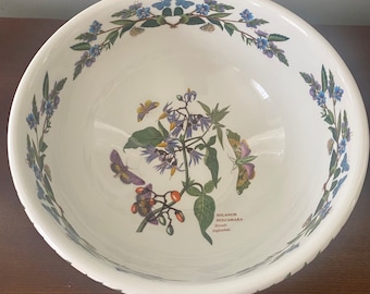 May include: A white ceramic bowl with a floral design featuring purple flowers, green leaves, and butterflies. The bowl has a rim with a repeating pattern of small blue flowers. The text "SOLANUM DULCAMARA Woody Nightshade" is printed on the bottom of the bowl.