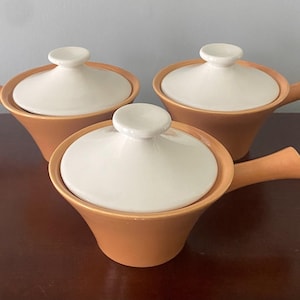 Könnte beinhalten: Drei Keramik-Serviergerichte mit Deckeln. Die Gerichte sind hellbraun und die Deckel sind weiß. Die Gerichte haben eine abgerundete Form und einen Griff auf einer Seite.