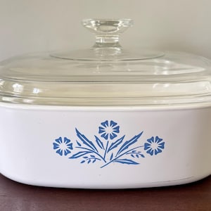 Puede incluir: Cazuela blanca con tapa de cristal transparente. El plato presenta un diseño floral azul. La tapa tiene un pomo de cristal transparente. La fuente rectangular tiene asas. Aproximadamente 25 cm de largo.