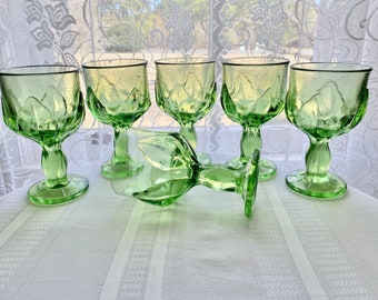 May include: Set of six green glass goblets with a textured design. The goblets are arranged in a semi-circle on a white tablecloth.