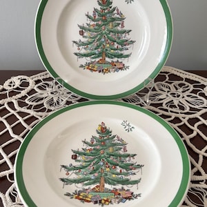 May include: Two white ceramic plates with a green border. Each plate features a detailed illustration of a Christmas tree decorated with ornaments and gifts. A small floral design is on the upper right.