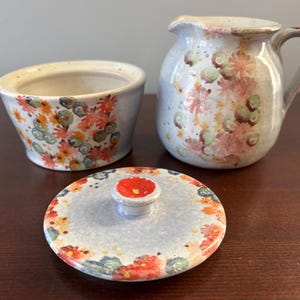 May include: A ceramic creamer and sugar bowl set with lids. The set is decorated with a floral pattern in shades of pink, orange, green, and blue on a white background.