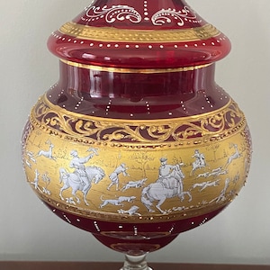 May include: A red glass decorative jar with gold accents and white painted hunting scene. The jar has a lid and sits on a clear glass pedestal with a gold rim.