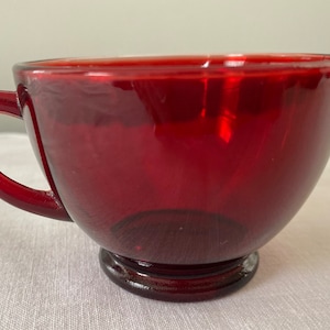 Könnte beinhalten: Eine rote Glas Tasse mit Henkel. Die Tasse hat einen abgerundeten Boden und einen ausgestellten Rand.