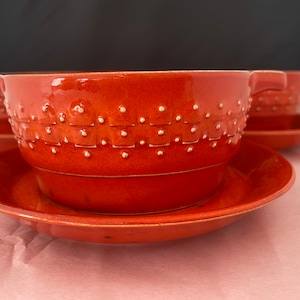 Puede incluir: Juego de cuatro cuencos de sopa de color terracota con platillos a juego. Cada cuenco tiene dos asas y un patrón texturizado con puntos blancos. Los cuencos y platillos están colocados sobre una superficie rosa.