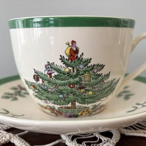 Puede incluir: Un juego de taza y platillo blancos con borde verde. La taza presenta un diseño de árbol de Navidad con Papá Noel y adornos. El platillo tiene un borde verde a juego e ilustraciones de acebo.