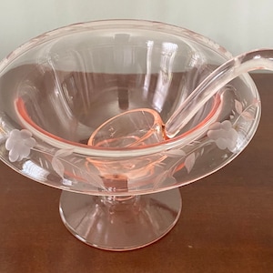 以下が含まれることがあります： ピンクの縁取りとエッチングされた花の模様が施された透明なガラス製のポンチボウル。ボウルは台座の上にあり、それに合うレードルが付いています。