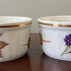 Puede incluir: Dos cuencos de cerámica blanca con borde dorado y diseño de mora. Los cuencos presentan un racimo de moras moradas y rojas con hojas verdes. Los cuencos están sobre una superficie marrón.