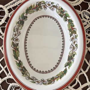 May include: Oval serving dish with a red border and a gold rim. The white dish features a floral and holly design around the edge and a decorative inner border. The dish is likely made of porcelain or ceramic.
