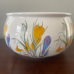 May include: White ceramic planter with a floral design featuring yellow, purple, and white flowers. The flowers are depicted in a realistic style and are arranged in a symmetrical pattern around the planter.