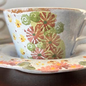May include: A white teacup and saucer with a floral pattern in shades of green, yellow, orange, and red. The cup has a handle and the saucer has a scalloped edge.