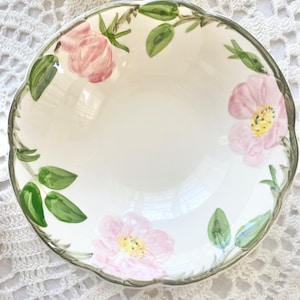 May include: A white ceramic bowl with a scalloped rim features a floral design of pink roses and green leaves. The bowl is perfect for serving snacks or desserts.