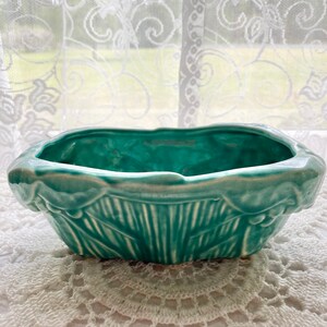 Pode incluir: Um vaso de cerâmica verde-azulado com uma borda festonada e um design em relevo de folhas e flores. O vaso está sobre uma renda branca de crochê.
