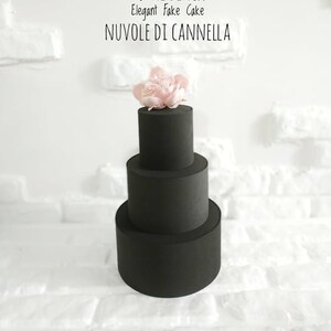 Könnte beinhalten: Ein dreistöckiger schwarzer Kuchen-Dummy mit einer rosa Blume oben. Der Kuchen ist mit "TOTAL BLACK Elegant Fake Cake NUVOLE DI CANNELLA" beschriftet.
