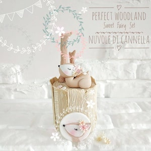 May include: A woodland-themed cake decorated with a sleeping fawn figurine and a fawn face on the side. The cake is decorated with a woodgrain pattern and pink flowers. The text "PERFECT WOODLAND Sweet Fairy Set NUVOLE DI CANNELLA" is written on the background.