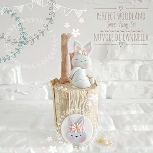 May include: A white bunny cake topper with a pink flower crown sits on a brown tree trunk cake. The cake is decorated with a white bunny face on a round brown circle. The text "PERFECT WOODLAND Sweet Fairy Set NUVOLE DI CANNELLA" is written on a white brick wall background.