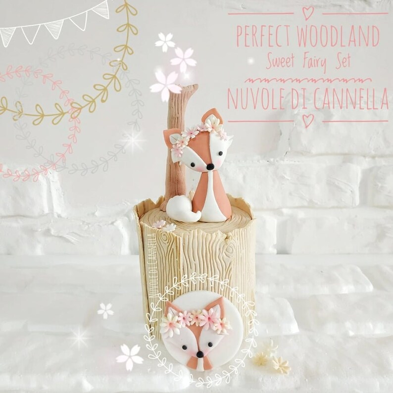 May include: A woodland themed cake topper featuring a small, orange fox with pink flowers around its head. The fox is sitting on a brown, tree trunk shaped cake stand. The cake topper is decorated with the text "PERFECT WOODLAND Sweet Fairy Set NUVOLE DI CANNELLA".