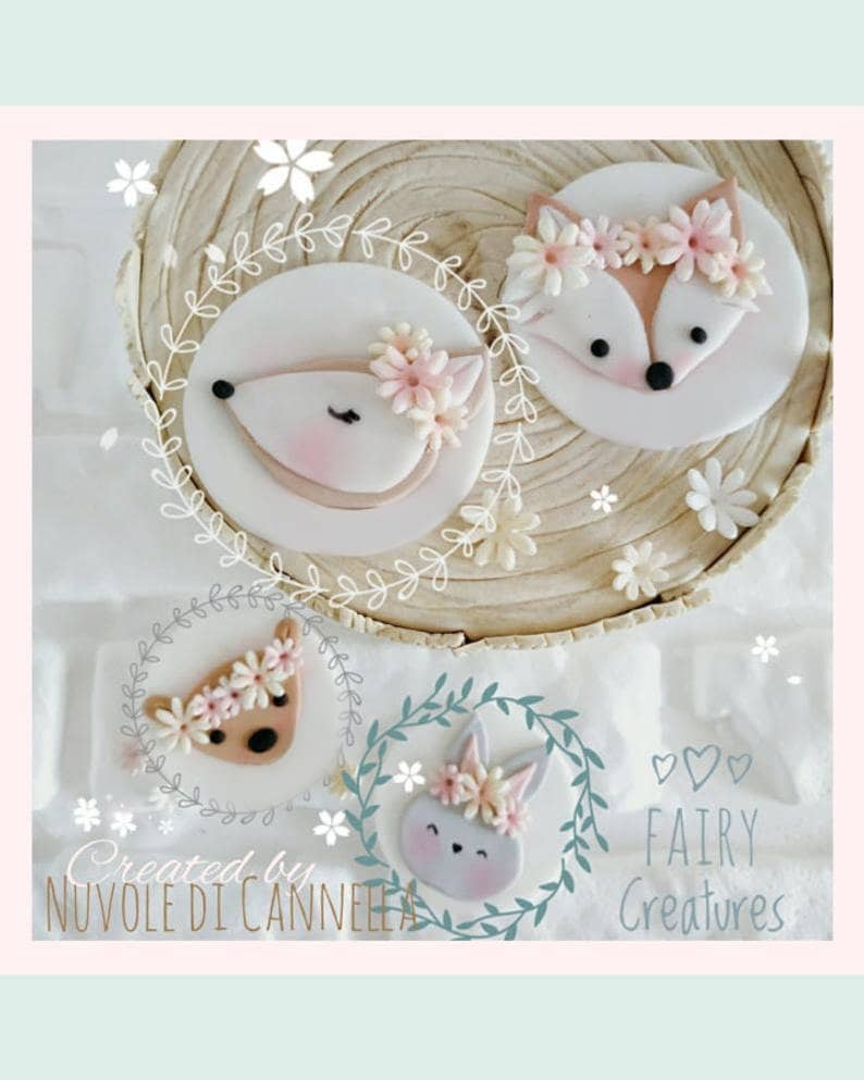 May include: Three fondant toppers featuring a fox, a deer, and a bunny. Each topper has a floral crown and is decorated in pastel pink, brown, and gray. The toppers are on a wooden background with the text "Created by Nuvole di Cannella Fairy Creatures".