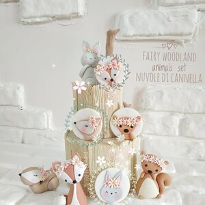 May include: A tiered cake decorated with fondant woodland animals. The cake is decorated with a woodgrain pattern and has a brown branch on top. The animals include a bunny, fox, squirrel, and deer. The cake is decorated with pink flowers and green leaves. The text "FAIRY WOODLAND animals set NUVOLE DI CANNELLA" is written on the cake.