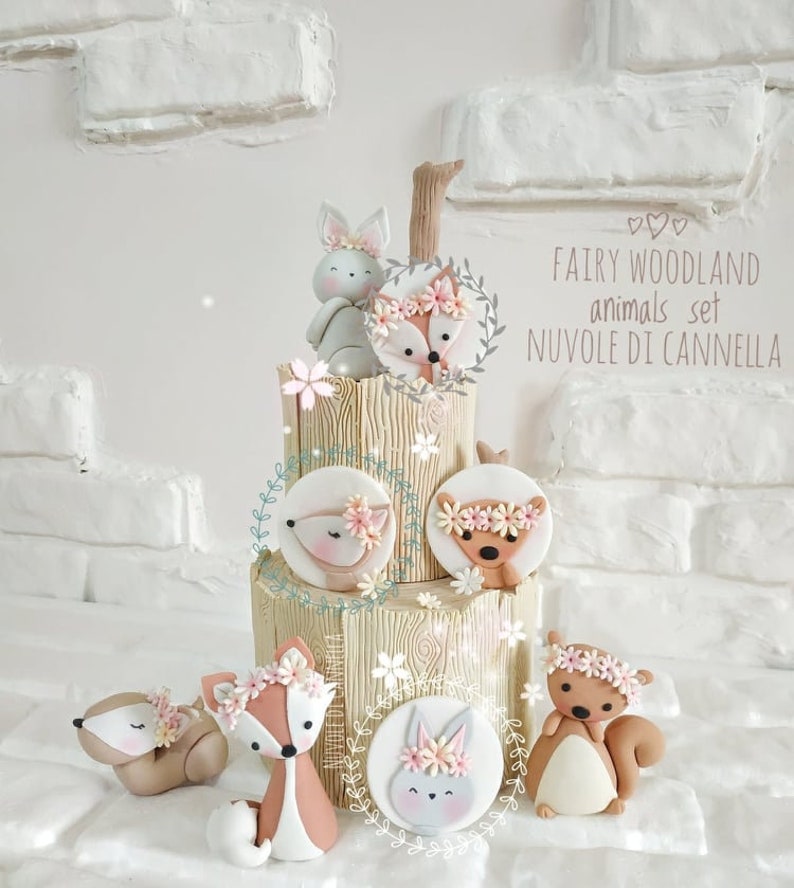 May include: A tiered cake decorated with fondant woodland animals. The cake is decorated with a woodgrain pattern and has a brown branch on top. The animals include a bunny, fox, squirrel, and deer. The cake is decorated with pink flowers and green leaves. The text "FAIRY WOODLAND animals set NUVOLE DI CANNELLA" is written on the cake.