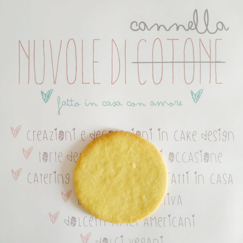 May include: A close-up of a light yellow cookie on a white background with the text "cannella NUVOLE DI COTONE" and "fatto in casa con amore" in pink and gray lettering.