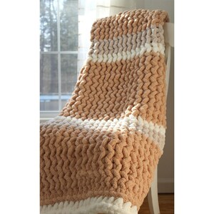 May include: A chunky knit blanket in shades of tan and white, draped over a white chair. The blanket features thick, textured yarn in alternating stripes of tan and white, creating a cozy and inviting aesthetic. The soft, plush material suggests warmth and comfort.