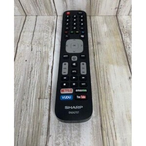 May include: A black Sharp EN2A27ST remote control with multiple buttons, including Netflix, Vudu, and YouTube. The remote has a directional pad and volume controls. The background is a light wood grain.