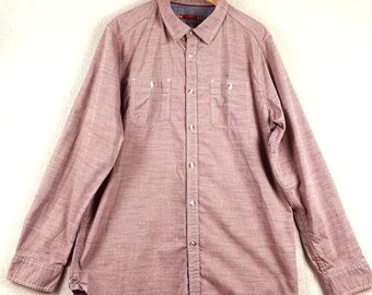 May include: A long-sleeved button-down shirt in a muted red-brown hue, hanging on a wooden hanger. The shirt features two chest pockets and a classic collar. The fabric has a subtle textured appearance.