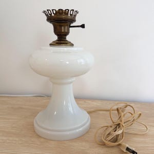 May include: A white milk glass table lamp base with a rounded body and a flared pedestal foot. The top features a brass-colored metal socket assembly with a small black switch. A beige electrical cord with a plug is coiled beside the lamp base.