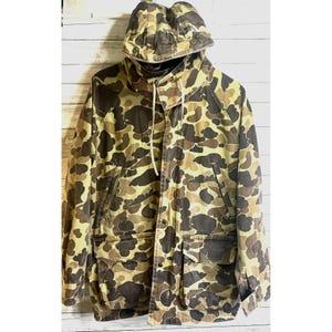 May include: A vintage camouflage jacket with a hood. The jacket features a pattern of brown, tan, and olive green shapes. It has a drawstring hood, multiple pockets, and a button-up front. The jacket is made of a durable fabric.