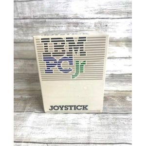 May include: A vintage IBM PCjr joystick box. The box is off-white with horizontal gray stripes and the IBM PCjr logo in blue, green, and gray. The word "JOYSTICK" is printed in black at the bottom.