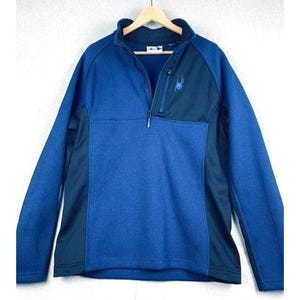 May include: A blue and black Spyder half-zip pullover. The top features a textured blue front panel, black side panels, and a black upper chest panel with a small spider logo. The jacket has a stand-up collar and a zipper closure.